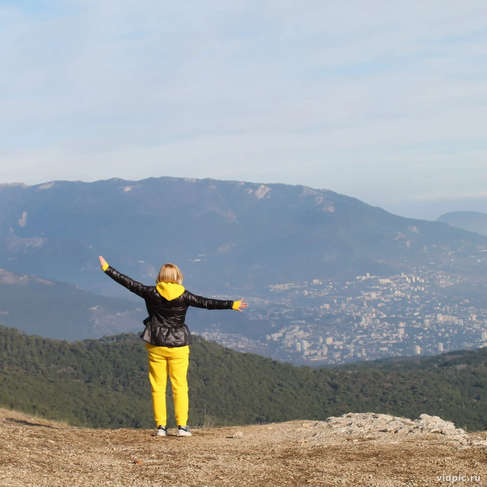 A girl in yellow pants and a black jacket stands on top of a mountain with her arms outstretched, enjoying the view of the mountain landscape and city below.