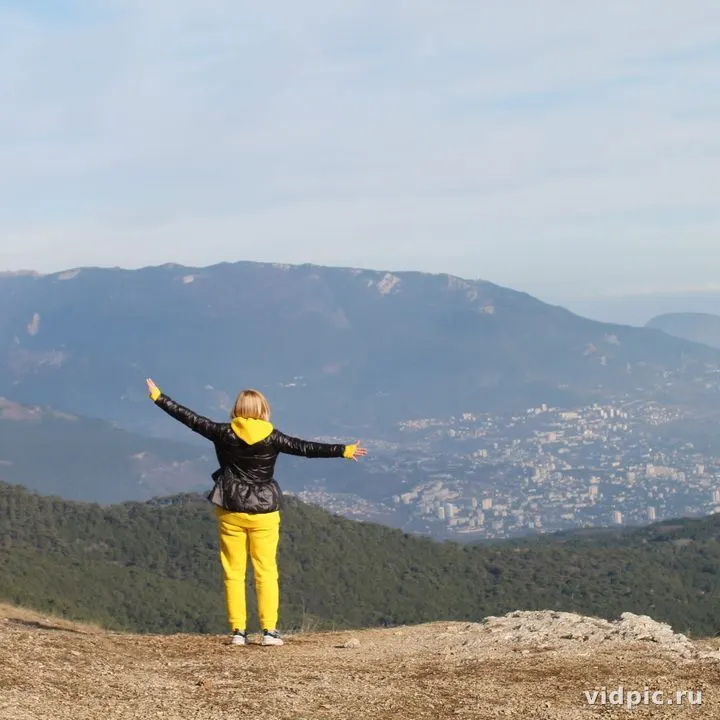 A girl in yellow pants and a black jacket stands on top of a mountain with her arms outstretched, enjoying the view of the mountain landscape and city below.