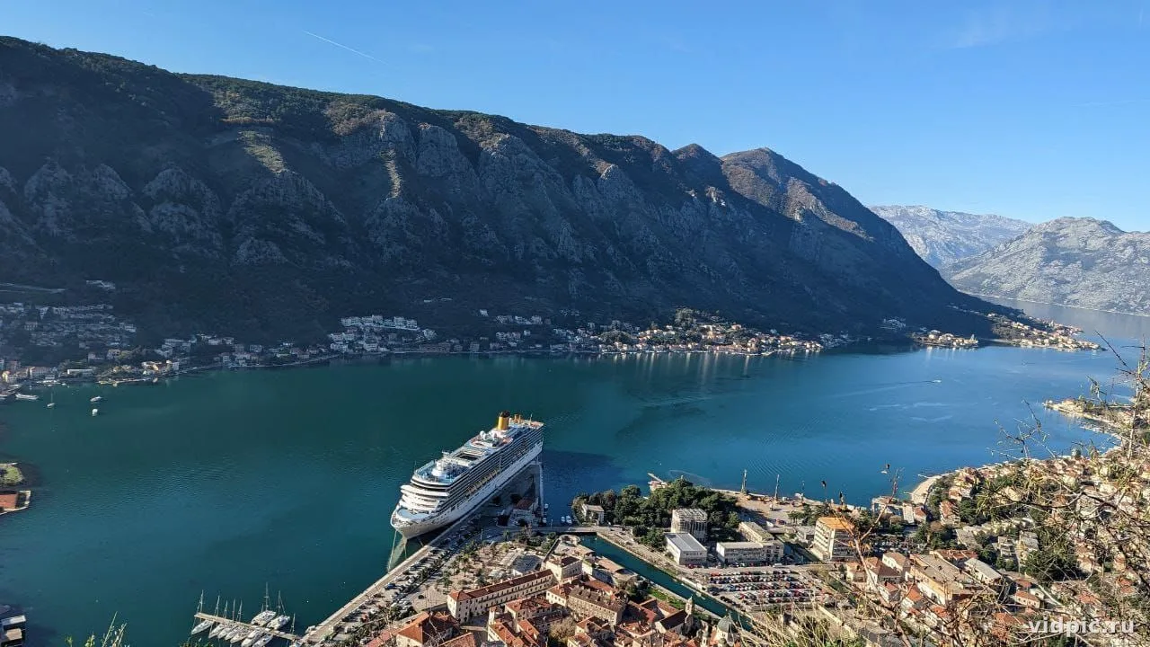 A city at the foot of mountains with a cruise ship. The blue of the water and sky contrasts with the white buildings and ship.