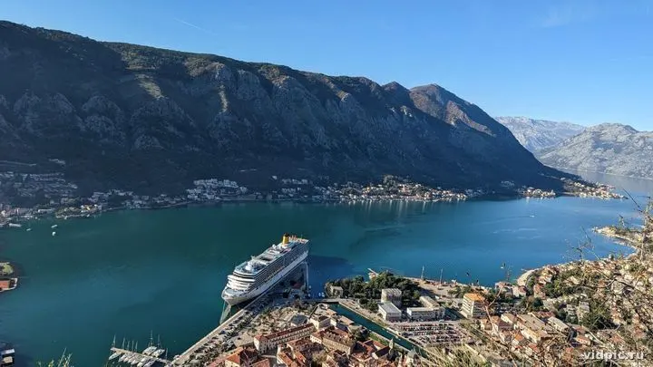A city at the foot of mountains with a cruise ship. The blue of the water and sky contrasts with the white buildings and ship.