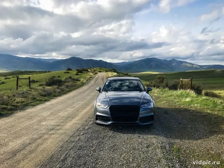 Gray car against the backdrop of mountains and cloudy sky. A gray car is parked on a dirt road leading through a green field towards mountains.